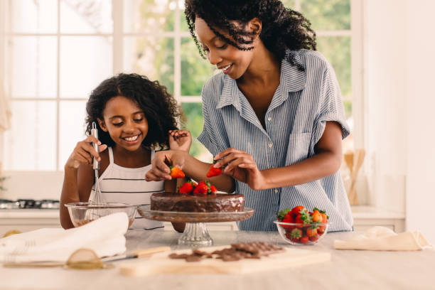 Woman with young girl making a cake at home Cute girl standing by her mother in kitchen making cake. Mother and happy daughter garnishing chocolate cake with fresh strawberries in kitchen. baking stock pictures, royalty-free photos & images
