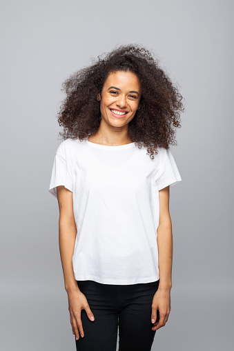 Portrait of beautiful young woman with afro hair wearing white t-shirt. Female student smiling at camera. Studio shot, grey background.