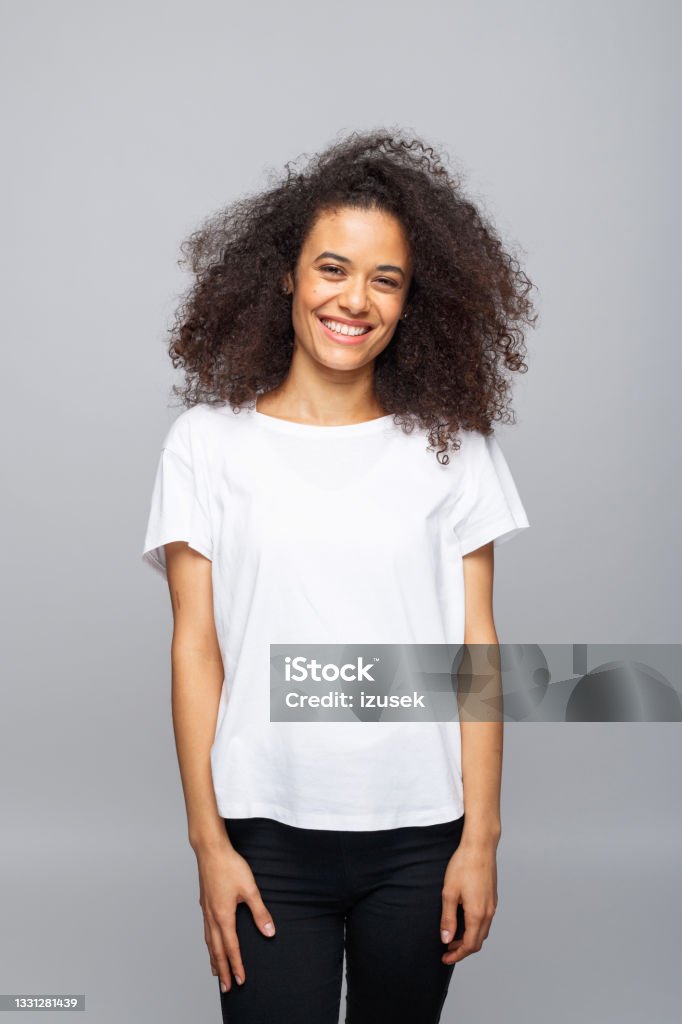 Cheerful young woman in white t-shirt Portrait of beautiful young woman with afro hair wearing white t-shirt. Female student smiling at camera. Studio shot, grey background. T-Shirt Stock Photo