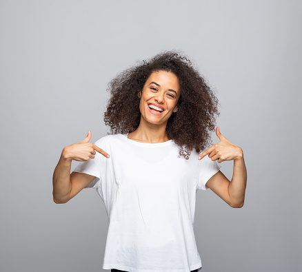 Portrait of beautiful young woman with afro hair wearing white t-shirt. Female student pointing with index fingers at t-shirt and smiling at camera. Studio shot, grey background.