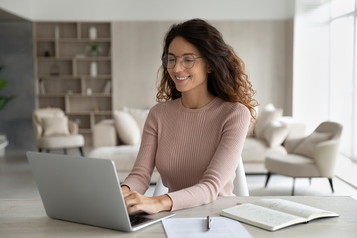 Happy millennial Latino woman in glasses sit at desk at home office look at laptop screen study online. Smiling young Hispanic female work type on computer, consult client customer distant on web.