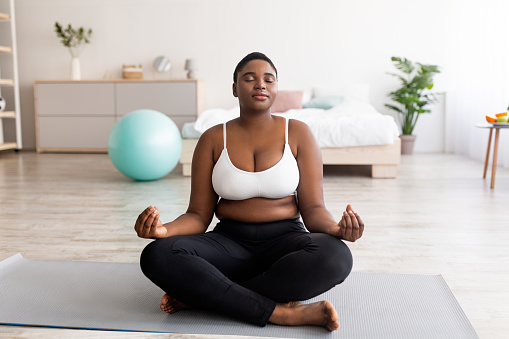 Plus size black woman sitting in lotus pose, meditating with closed eyes, practicing yoga at home. Overweight young lady doing breathing exercises. Wellness, mindfulness concept