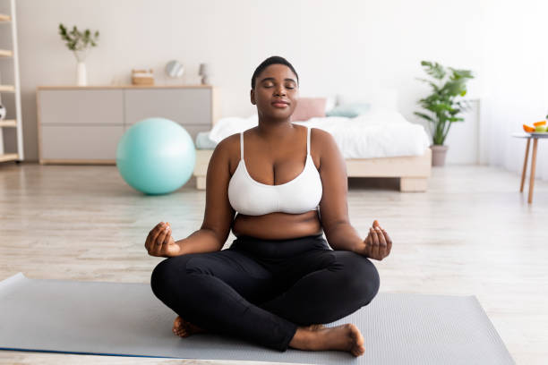 Plus size black woman sitting in lotus pose, meditating with closed eyes, practicing yoga at home Plus size black woman sitting in lotus pose, meditating with closed eyes, practicing yoga at home. Overweight young lady doing breathing exercises. Wellness, mindfulness concept voluptious stock pictures, royalty-free photos & images