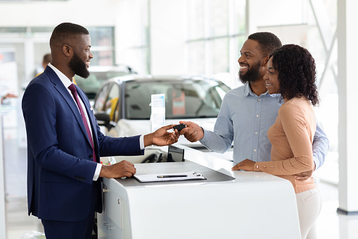 Car Sales. Handsome Black Manager Giving Key To Happy Couple In Dealership Center After Buying New Vehicle, Greeting African American Spouses With Successful Purchase In Automobile Showroom