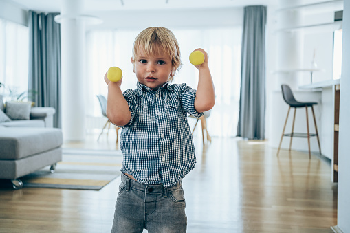 Cute little boy lifting weights in living room at home.