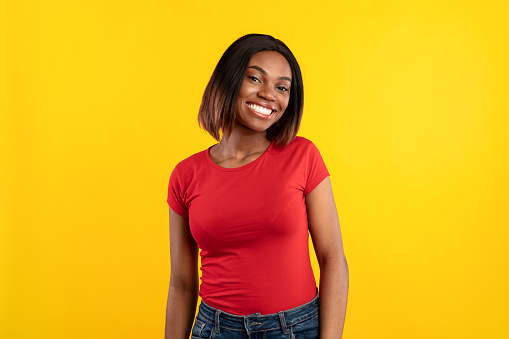 African American Lady Posing Smiling To Camera Standing Over Yellow Studio Background, Wearing Red T-Shirt. Portrait Of Cheerful Black Young Woman With Toothy Smile Concept