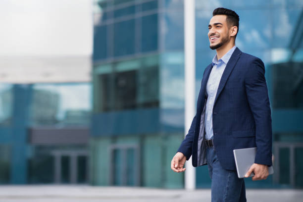 feliz joven empresario árabe caminando de la oficina, sosteniendo el bloc de notas - middle eastern ethnicity business men manager fotografías e imágenes de stock