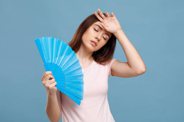 Upset millennial lady using hand fan over blue background Upset millennial lady using hand fan over blue studio background, touching her forehead, closing eyes. Young pretty woman cooling herself while summer heat, holding paper fan, closeup relieved face stock pictures, royalty-free photos & images