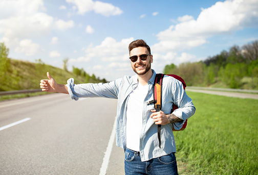 Handsome young man with backpack hitchhiking on road, showing thumb up gesture, stopping passing car. Autostop journey, summertime travel, freedom and tourism concept