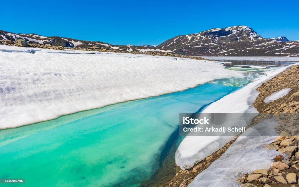 Frozen turquoise lake Vavatn panorama in summer landscape and mountains with snow in Hemsedal Norway. Frozen turquoise lake Vavatn panorama in summer landscape and mountains with snow and blue sky in Hemsedal Norway. Awe Stock Photo