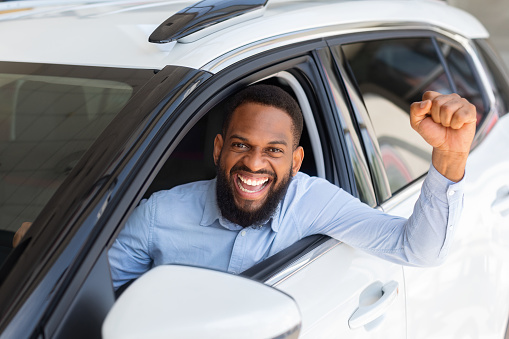 Excited Black Guy Posing In His New Car After Buying, Raising Fists And Exclaiming With Joy Through Window, Joyful African American Man Celebrating Purchasing Modern Vehicle In Dealership Center