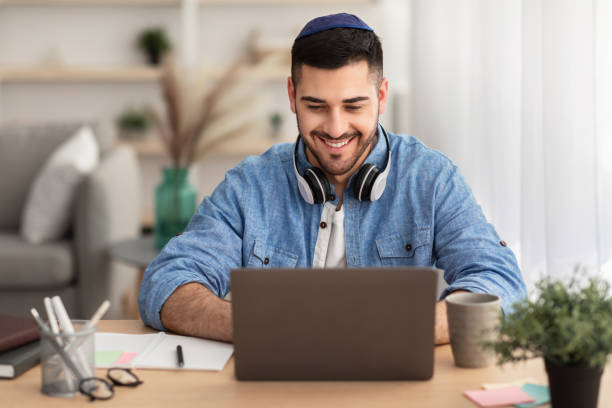 Smiling israeli man working on laptop at home Business And Entrepreneurship. Happy Jewish Man In Kippa Working On Laptop Wearing Headphones On Neck Sitting At Workplace In Modern Home Office. Independent Contractor Enjoying Distance Job yarmulke photos stock pictures, royalty-free photos & images