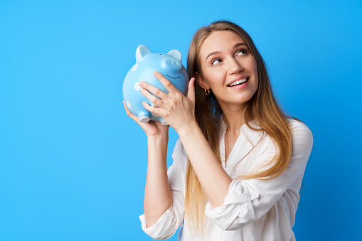 Beautiful smiling young woman with piggy bank on blue background, close up