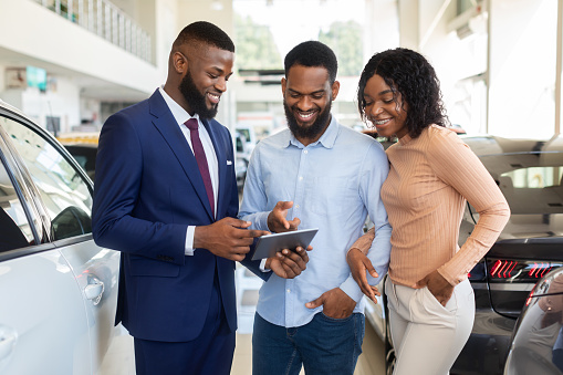 Handsome Car Seller Showing Purchasing Details On Digital Tablet To Young Black Couple In Dealership Center, Happy African American Spouses Buying New Vehicle In Automobile Showroom, Closeup