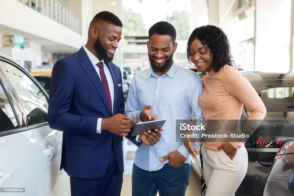 Handsome Car Seller Showing Purchasing Details On Digital Tablet To Black Couple Handsome Car Seller Showing Purchasing Details On Digital Tablet To Young Black Couple In Dealership Center, Happy African American Spouses Buying New Vehicle In Automobile Showroom, Closeup Car Dealership Stock Photo
