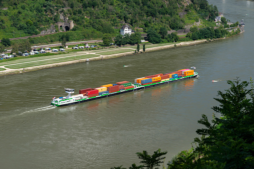Loreley, Germany - 25 July 2021. A large barge carrying a lot of containers on the Rhine River in western Germany, visible hills overgrown with trees.