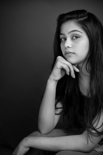 Black and white close-up portrait of young woman sitting against dark background looking away at looking at the camera with a blank expression and contemplating.