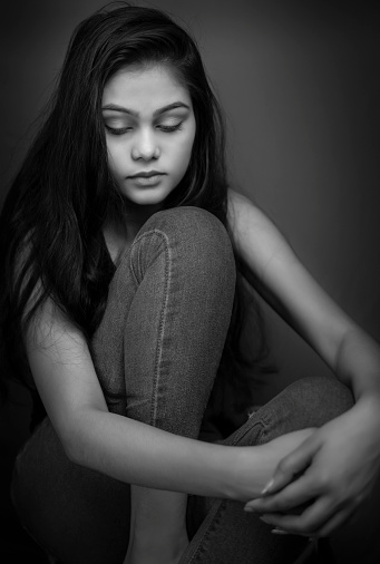 Black and white close-up portrait of sad, tensed young woman looking away and contemplating with a blank expression against a dark background.