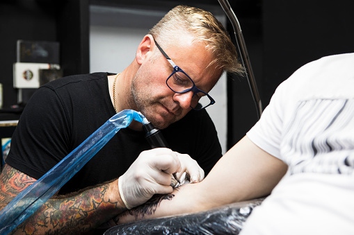 Tattoo artist working in a tattoo studio. He is wearing a black t-shirt and glasses