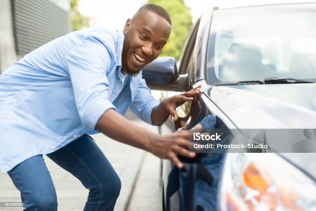 Happy African American Guy Touching His New Auto Happy Car Buyer, New Car Owner Concept. Portrait Of Excited Young African American Guy Touching His New Luxury Automobile After Purchase In Dealership Showroom. Selective Focus Car Stock Photo