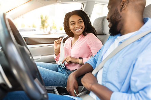 Safety First. Smiling African American Couple Fastening Seatbelts Before Driving In City, Sitting Inside New Luxury Car, Following Safe Ride Rules, Ready For Travel With Auto, Looking At Each Other