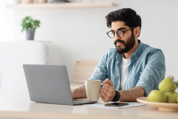 istock Modern worker in self-isolation. Concentrated arab man drinking coffee and working on laptop at table in kitchen 1331296232