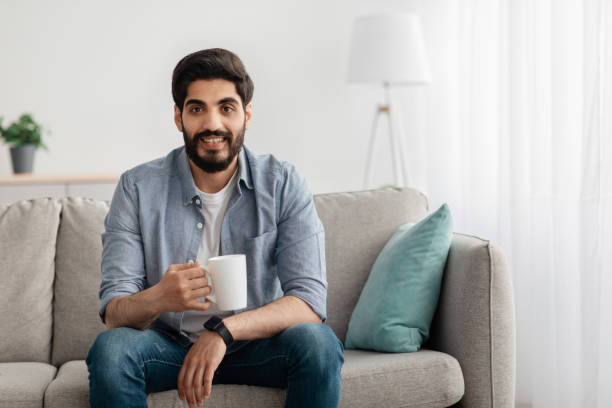 istock Young relaxed arab guy sitting on couch and drinking tea or coffee, resting in living room interior, free space 1331297498