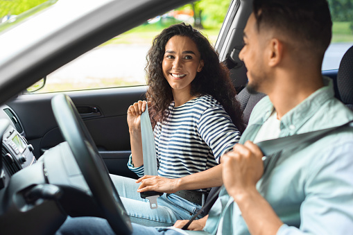 Excited millennial arab couple fastening seat belt, looking at each other and smiling, going vacation by car, side view, closeup. Mixed race young family having weekend trip, enjoying nice weather