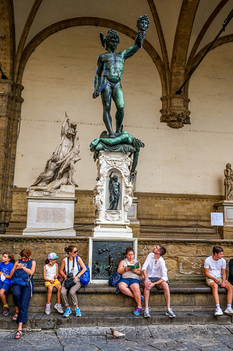 Florence, Tuscany, Italy, August 11 -- Some tourists rest under the magnificent bronze sculpture of Perseus who raises the head of Medusa after cutting it, made by Benvenuto Cellini and preserved in the Loggia Dei Lanzi, in Piazza della Signoria, in the historic heart of Florence near the Uffizi palace. Commissioned by Cosimo de Medici, the statue represents the cutting of the republican experiences of the city and of political divisions. The Loggia dei Lanzi, an open structure with arch and columns designed by the architects Benci and Simone Talenti in 1376 on the right side of Piazza della Signoria, was used for the official ceremonies of the Florentine government. Since 1982 the historic center of Florence has been declared a World Heritage Site by Unesco. Image in high definition format.