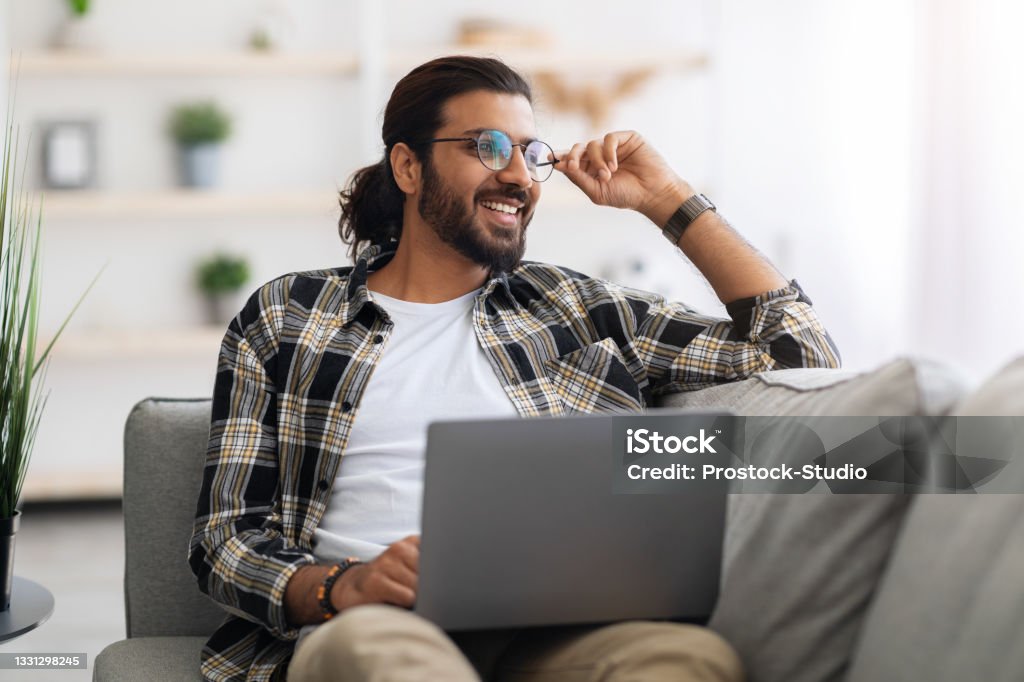 Dreamy arab guy freelancer working from home, using laptop Dreamy arab guy freelancer working from home, wearing glasses, sitting on couch, using laptop and looking at copy space, thinking about career opportunities or looking for creative solution Computer Programmer Stock Photo