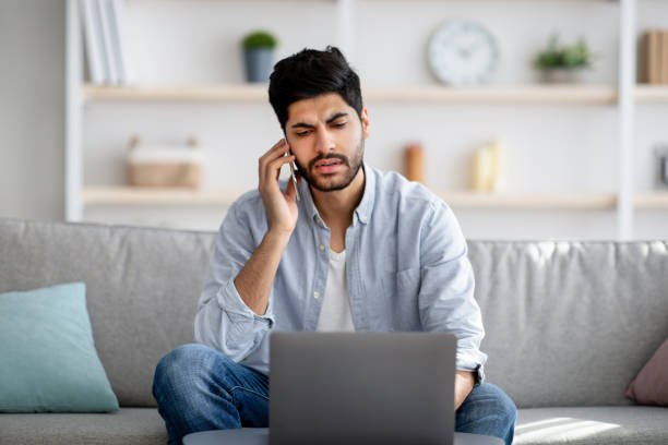 istock Remote business. Concerned arab freelancer talking on cellphone and using laptop computer, sitting on sofa at home 1331298458