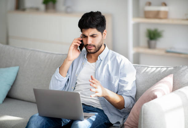 istock Computer problems. Displeased arab man talking on smartphone and looking on laptop screen at home 1331299188