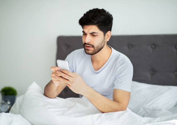 istock Arab man using smartphone, browsing internet and texting on phone sitting on bed at home, free space 1331299803
