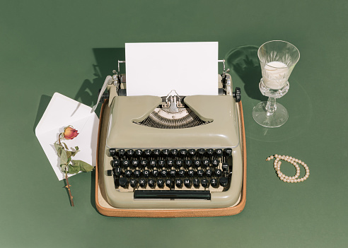 Retro styled scene with a typewriter, white letter, drinking glass, dry rose flower, and pearl bracelet with strong shadows against a green background. Minimal vintage aesthetics. Art direction.