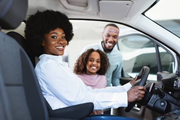 giro di prova. donna di colore seduta sul sedile del conducente di un'auto nuova, scegliendo il veicolo con marito e figlia in concessionaria - car african descent shopping car dealership foto e immagini stock