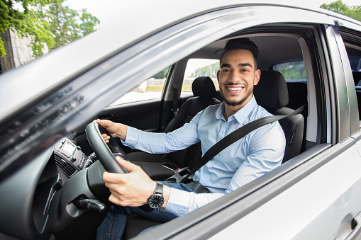Cheerful arab businessman driving car, shot from outside, going to office or business trip, copy space. Happy middle-eastern guy in formal outfit with fasten seat belt driving his brand new car