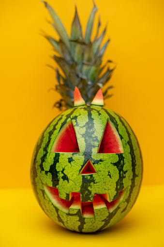 Pineapple hiding behind watermelon. Carved Halloween green jack-o'-lantern. Alternative Halloween pumpkin as a watermelon and pineapple against yellow  wall with space for text. Summerween time.