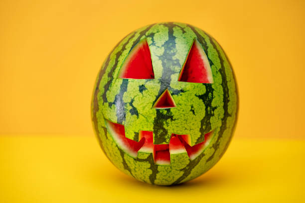 Watermelon with a smiling face like a pumpkin for Halloween. Tropical jack-o-lantern watermelon on yellow background. Summerween time. Carved Halloween green jack-o'-lantern. Alternative Halloween pumpkin as a watermelon on yellow wall with space for text. Watermelon with a smiling face like a pumpkin for Halloween. fruit carving stock pictures, royalty-free photos & images