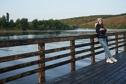 Full length portrait of young woman in casual warm outfit posing near lake. Pretty lady with brown hair leaning on handrails on wooden pier.