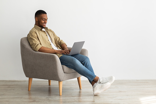 Side-View Of Black Freelancer Man Using Laptop Computer Working Online And Browsing Internet Sitting In Armchair Over Gray Wall Indoors. Internet And Technology, People And Gadgets Concept