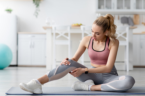 Training injury at home. Woman feeling pain after exercising at home. Desperate young blonde lady in sportswear suffering from knee pain, in minimalist living room, kitchen interior, copy space