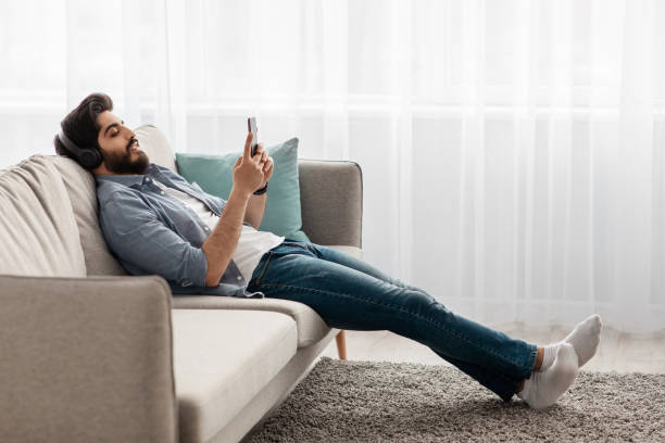 istock Domestic relax. Young arab guy with wireless headphones and smartphone resting on comfortable sofa at home, side view 1331304079