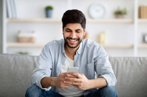 istock Handsome smiling arab man sitting with smartphone in hands and messaging with friends, relaxing with cellphone on couch 1331305073
