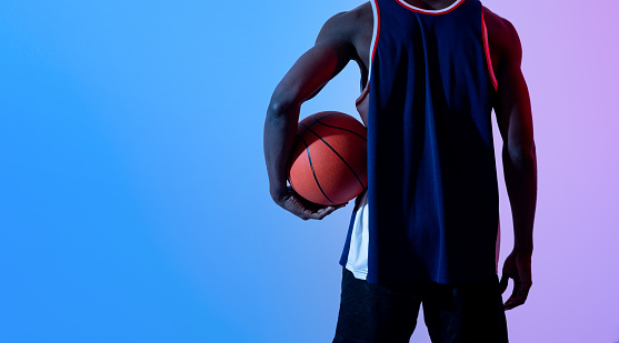 Unrecognizable black basketball player holding ball in neon light, banner design with copy space. Young African American sportsman ready to participate in streetball match, play in tournament, closeup