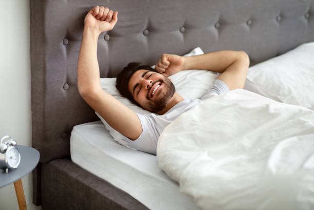 istock Rested arab man waking up and stretching hands, wellslept guy lying in bed, awakening in cozy bedroom and smiling 1331305132