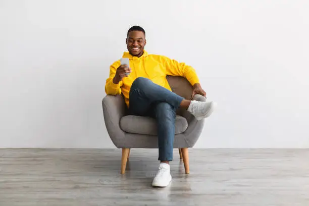 Photo of Black Man Using Smartphone Sitting In Armchair Near Gray Wall