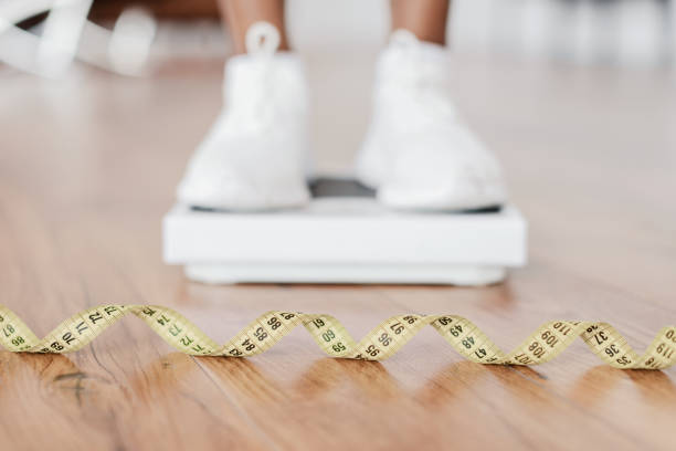 Closeup Black Woman Standing On Scales, Focus On Measure Tape Weight Control Concept. Selective focus on yellow measuring tape on the floor and blurred view of african woman standing on scales, checking result of her slimming diet. Dieting, healthy living photos of anorexia pictures stock pictures, royalty-free photos & images