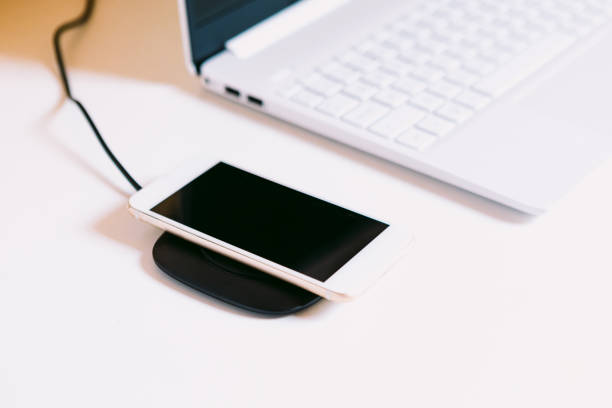 istock Hand with white mobile smart phone, charging on a cordless charger base next to a laptop on a white work table 1331309285