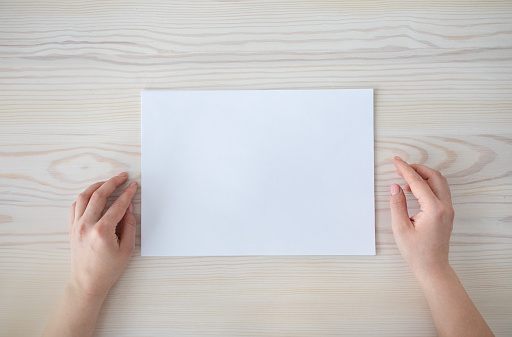 Female hands with white blank paper sheet on wooden table background, top view, empty space, flat lay. Mockup for your design or advertisement