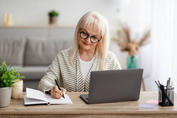 istock Mature lady writing in notebook and using laptop at home 1331311885